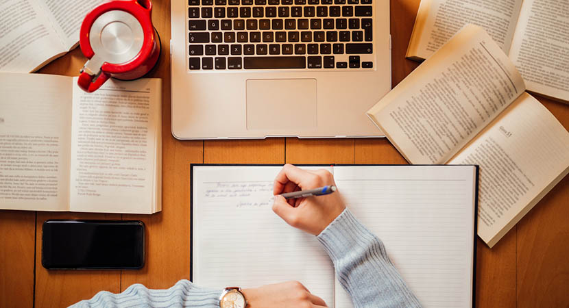 Person writing in a notebook surrounded by books and a computer