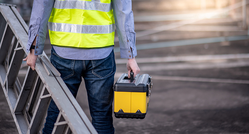 Maintenance worker man with safety helmet and green vest carrying aluminium step ladder and tool box at construction sit