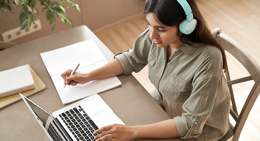 Female student wear headphones learning online watching webinar class looking at laptop computer elearning remote lesson making notes or video calling virtual conference meeting teacher at home.